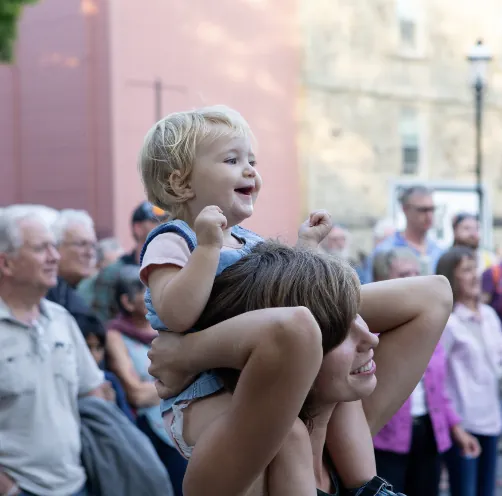 A grinning toddler on a parent’s shoulders in the crowd at a Harvest street show